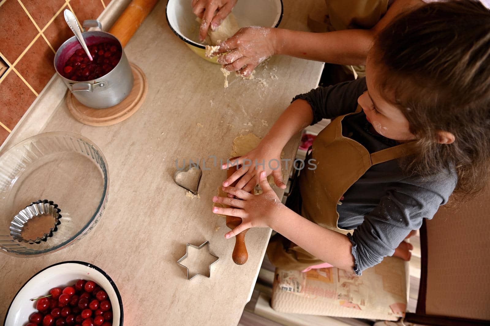 Overhead view of beautiful little girl, wearing a beige chef's apron, kneading and rolling dough with a wooden rolling pin. Cutting molds and fresh ingredients on a kitchen countertop. Baking concept
