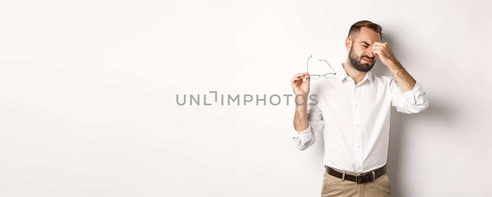 Tired businessman take-off glasses and rubbing eyes, standing exhausted against white background.