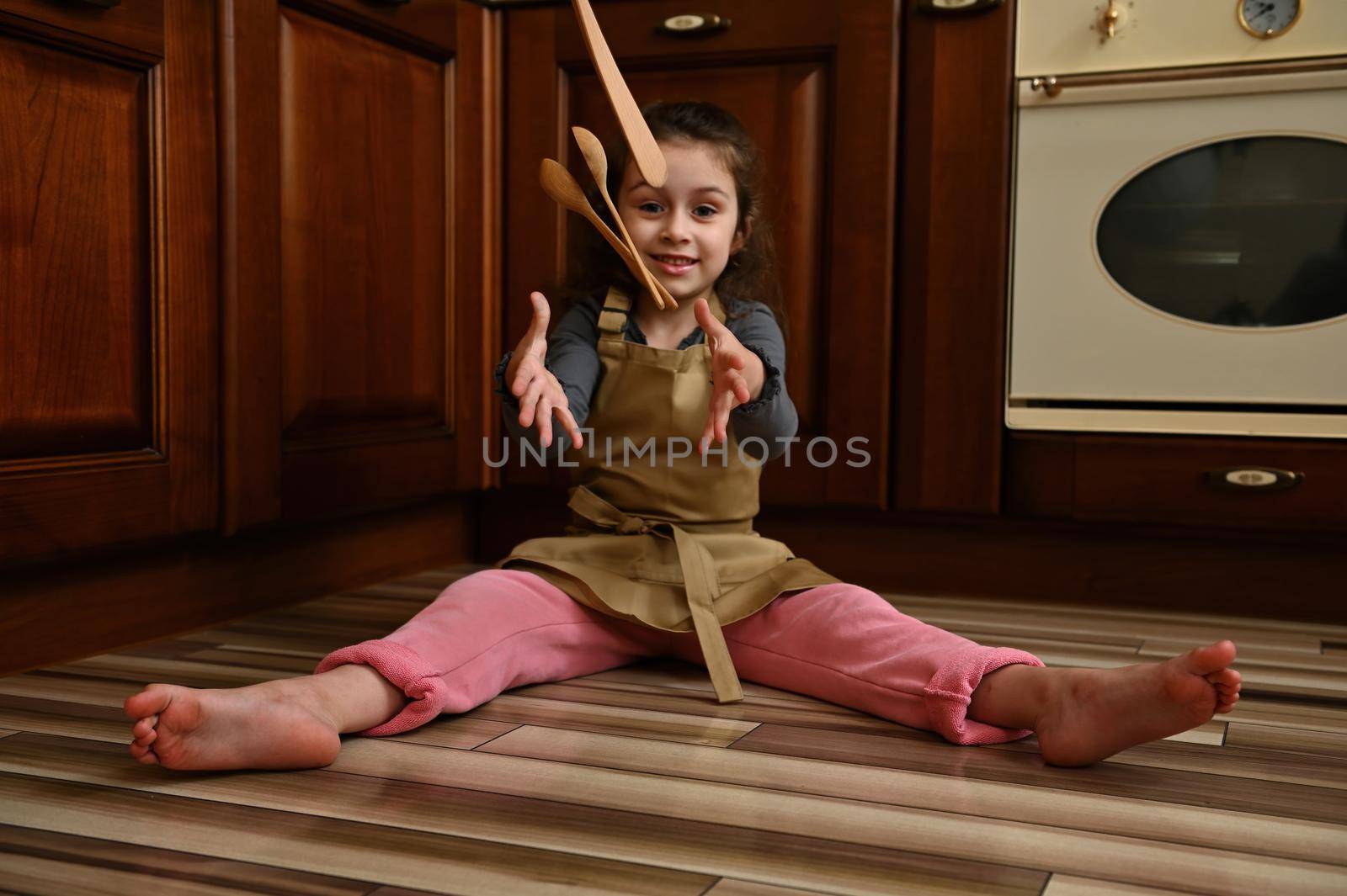 Selective focus on charming child, beautiful little girl, wearing beige chef apron, sitting barefoot on the floor by the oven, in the home rustic kitchen and throwing spoons smiling looking at camera