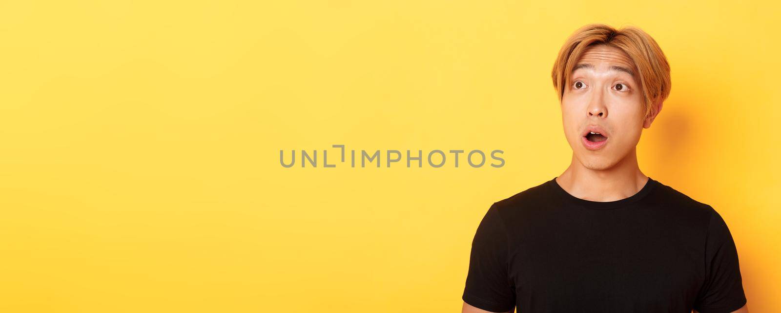 Close-up of impressed asian handsome guy, looking upper left corner with dropped jaw and amazement, standing over yellow background.