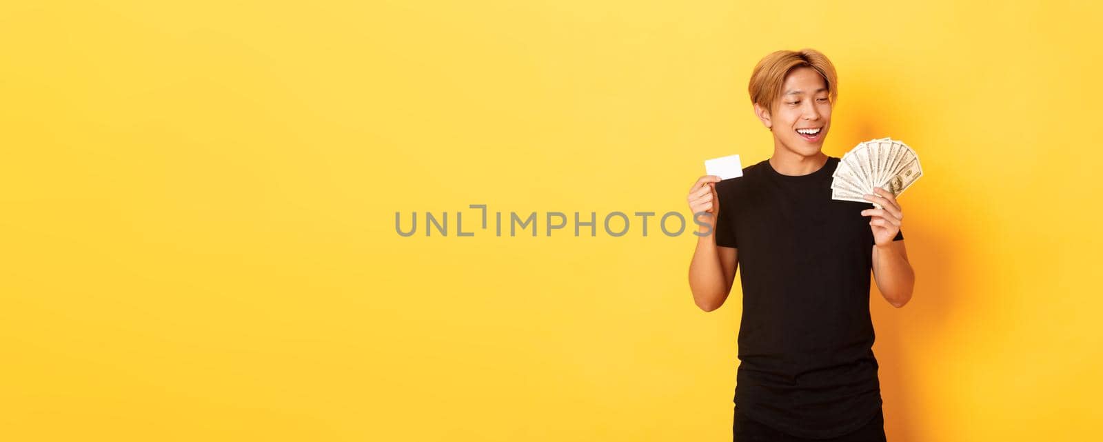 Happy and lucky asian guy looking pleased at money while showing credit card and cash, standing yellow background by Benzoix