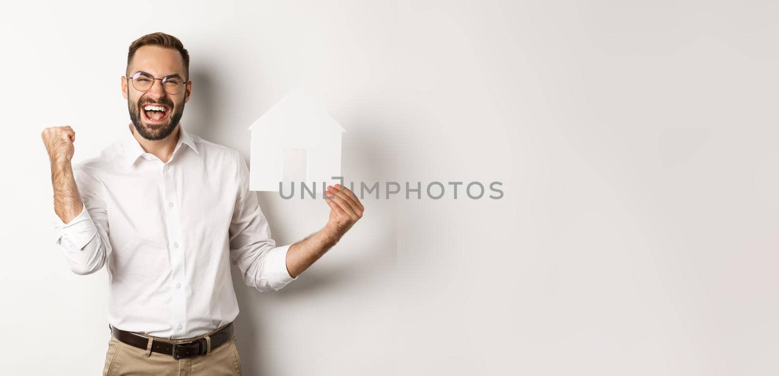 Real estate. Satisfied man rejoicing of founding perfect home apartment, holding paper house model, standing over white background by Benzoix