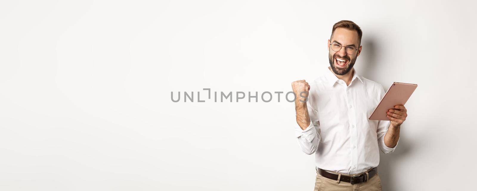 Successful businessman rejoicing on winning online, reading on digital tablet and making fist pump, triumphing, standing over white background.
