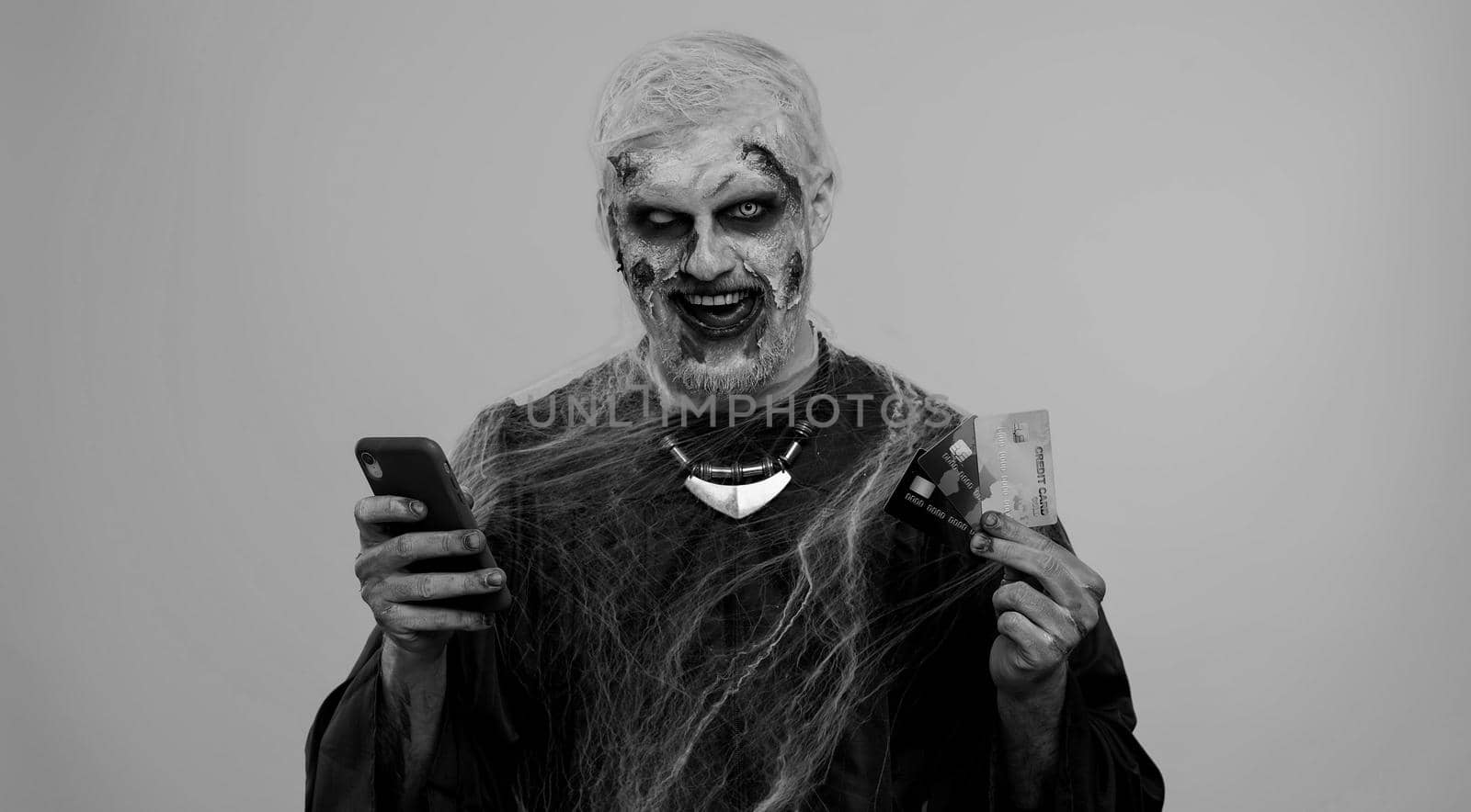 Sinister man with horrible scary Halloween zombie make-up using credit bank cards and mobile phone, transferring money purchases online shopping. Dead guy with wounded bloody scars face, gray room