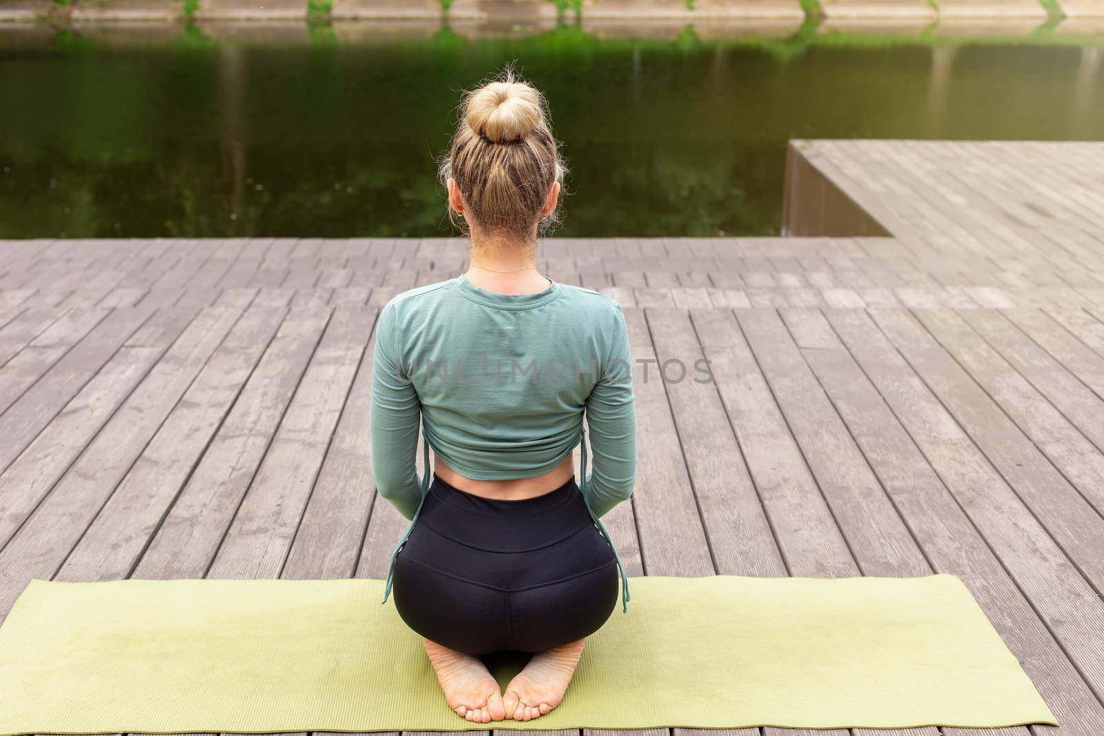 A slender woman in a green top and black leggings, sitting on a green sports mat, on a wooden platform by a pond in the park. Rear view. Copy space
