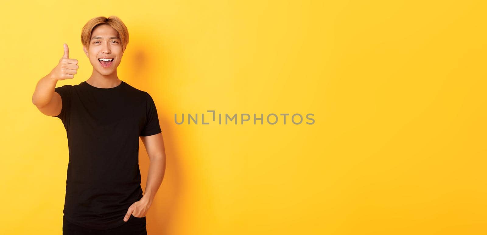 Portrait of pleased handsome asian guy, showing thumbs-up in approval, standing over yellow background by Benzoix