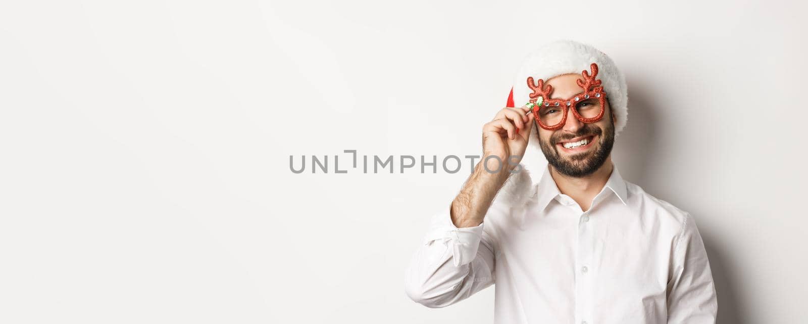 Close-up of handsome bearded guy in xmas party glasses and santa hat, smiling and wishing merry christmas, standing over white background by Benzoix