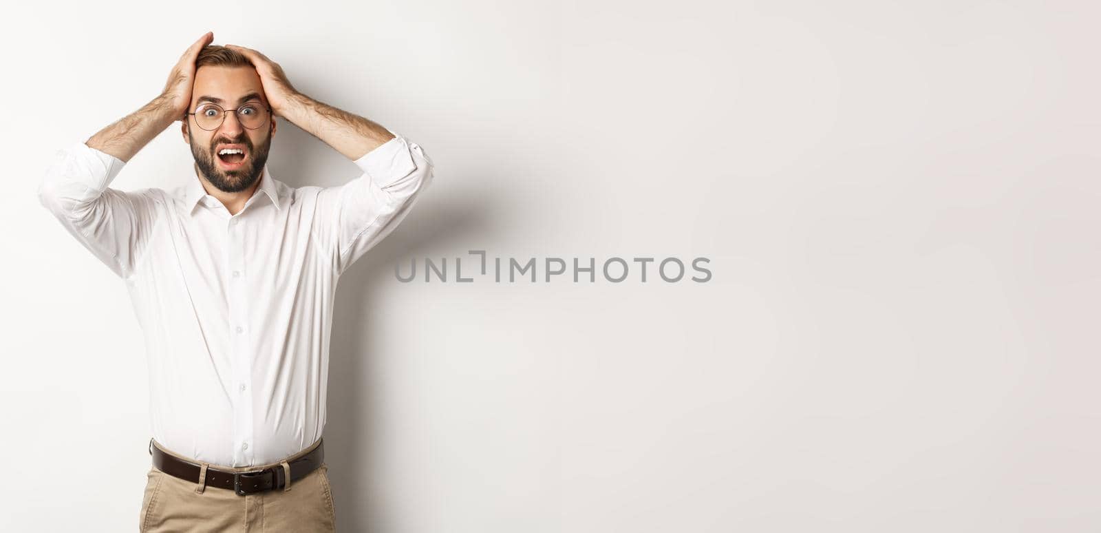 Frustrated businessman holding hands on head, looking shocked and anxious, standing over white background.