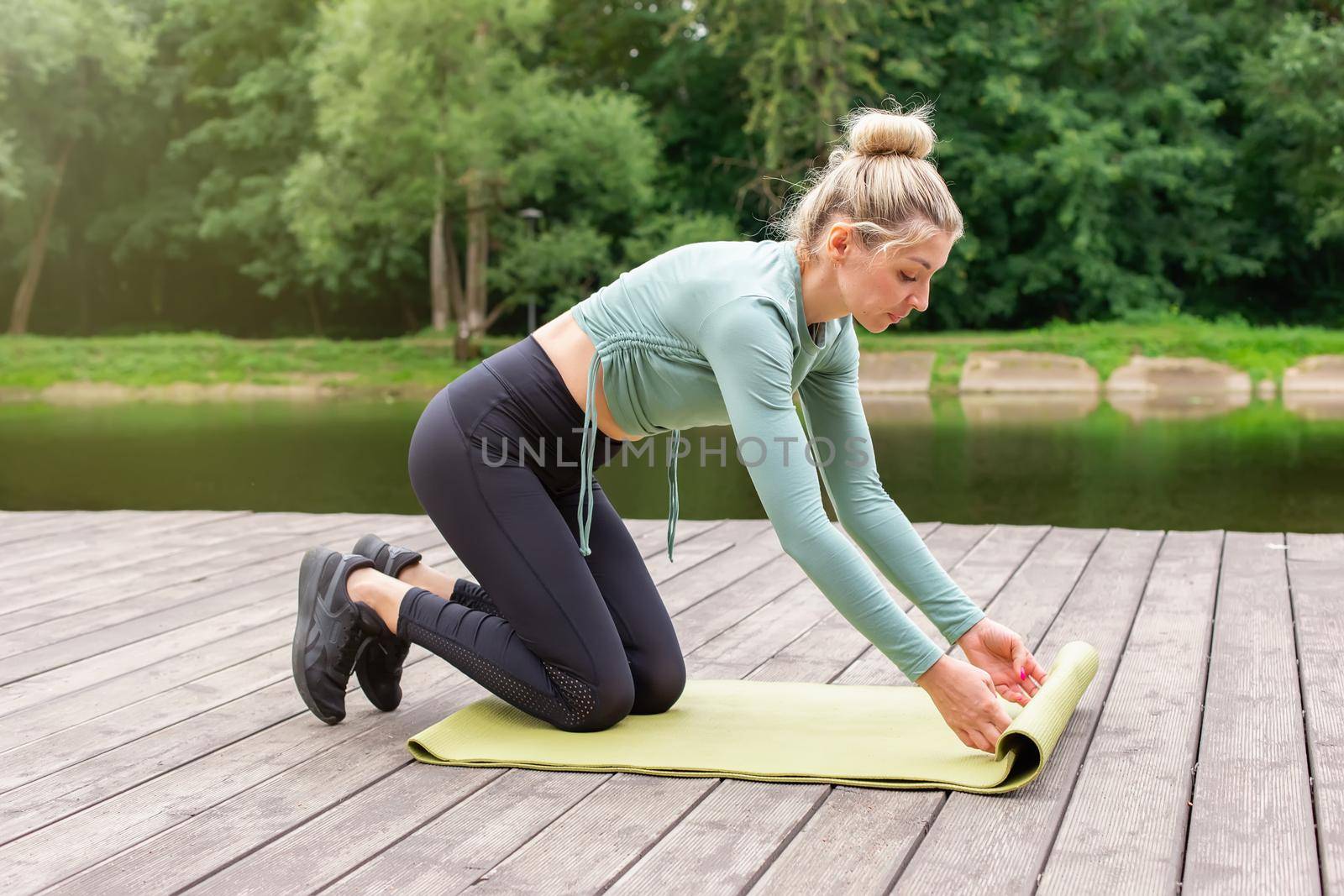 A woman in a green top and black leggings, in the summer by pond, unwinds a green gym mat, prepares for sports. side view. Copy space