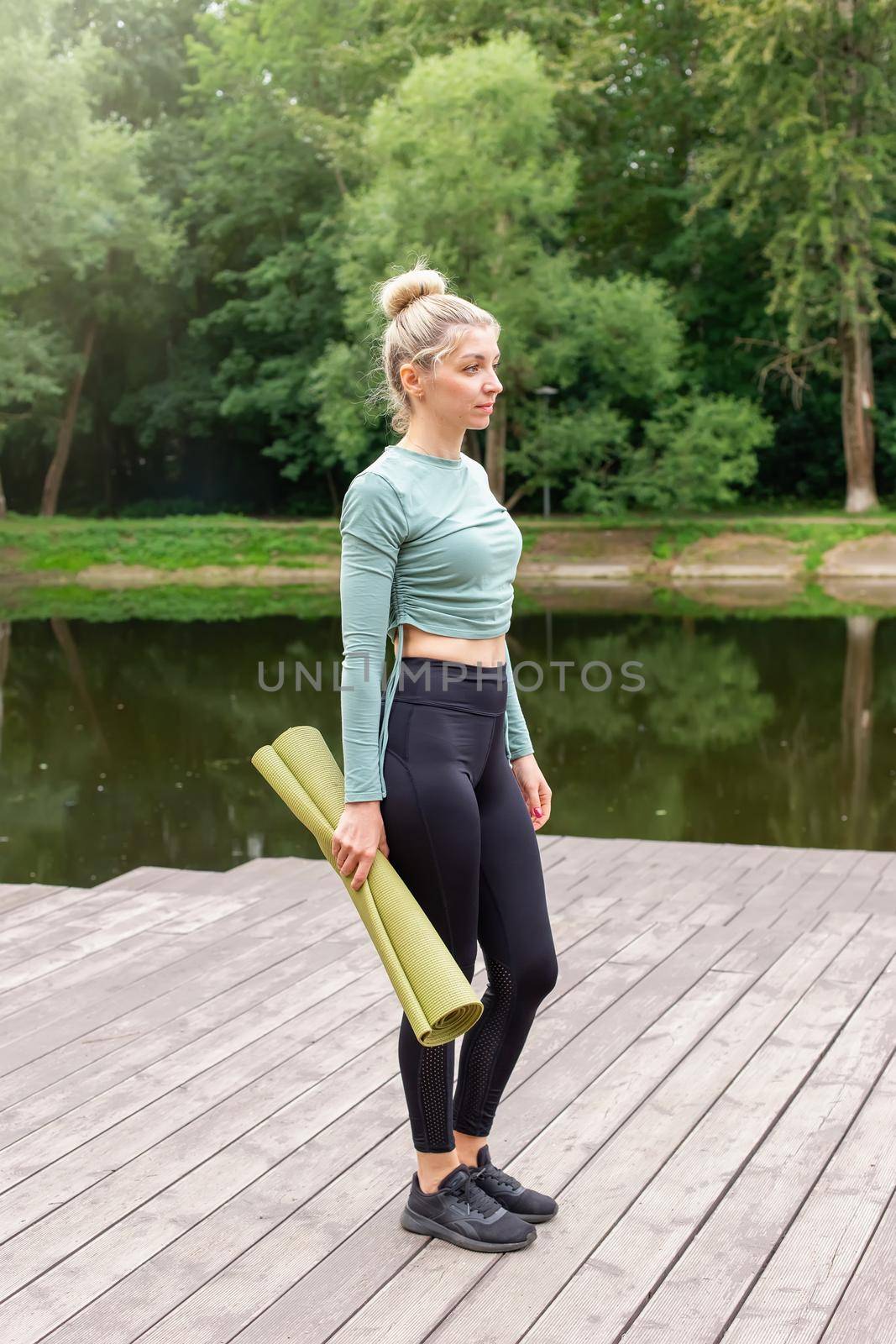 A beautiful woman in a green top and dark leggings, standing in summer, with a green gym mat for sports. by Zakharova