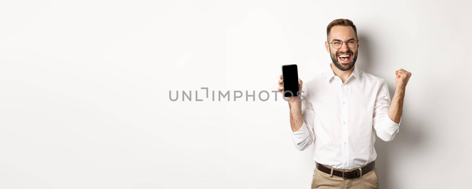 Successful business man showing mobile screen, rejoicing on winning online prize, achieve app goal, standing against white background.