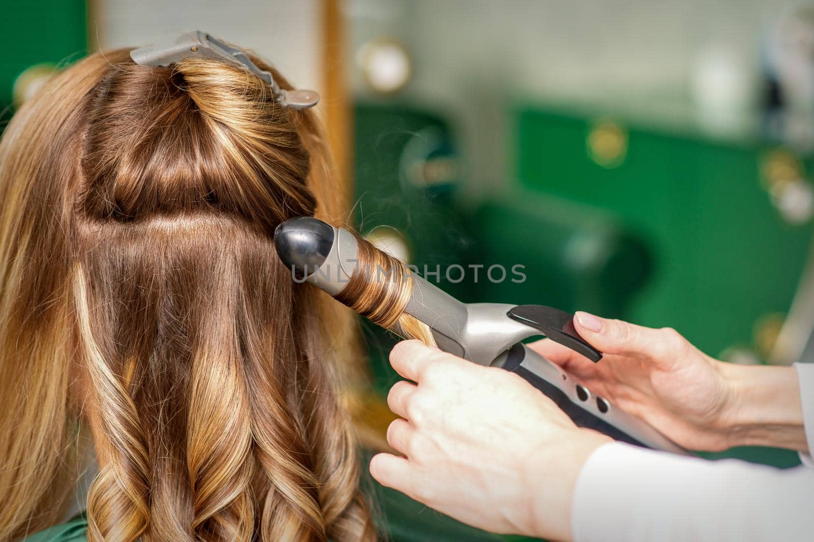 Creating curls with curling irons. Hairdresser makes a hairstyle for a young woman with long red hair in a beauty salon. by okskukuruza