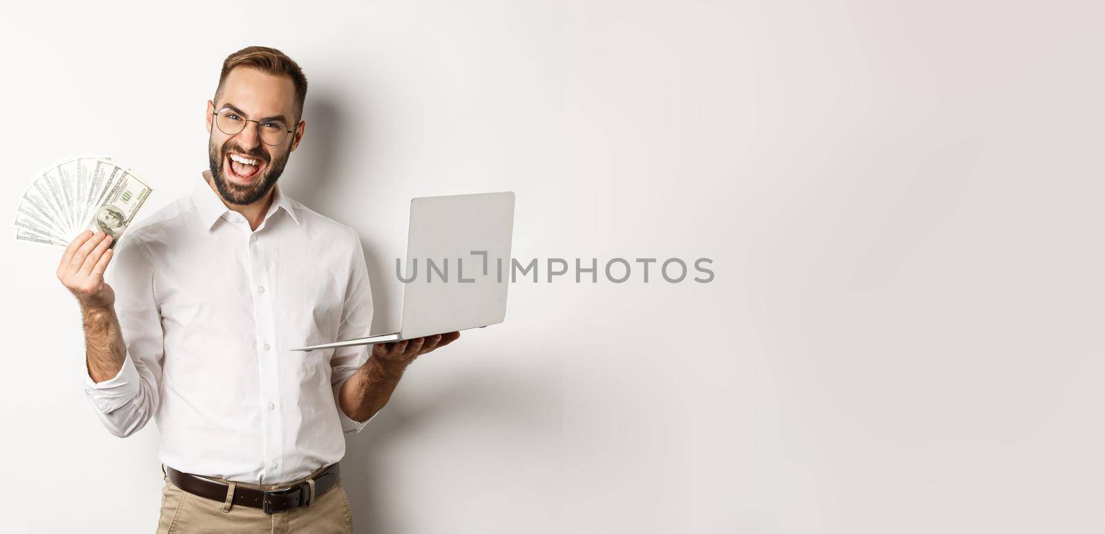 Business and e-commerce. Excited businessman holding money dollars and laptop, working online, standing over white background.