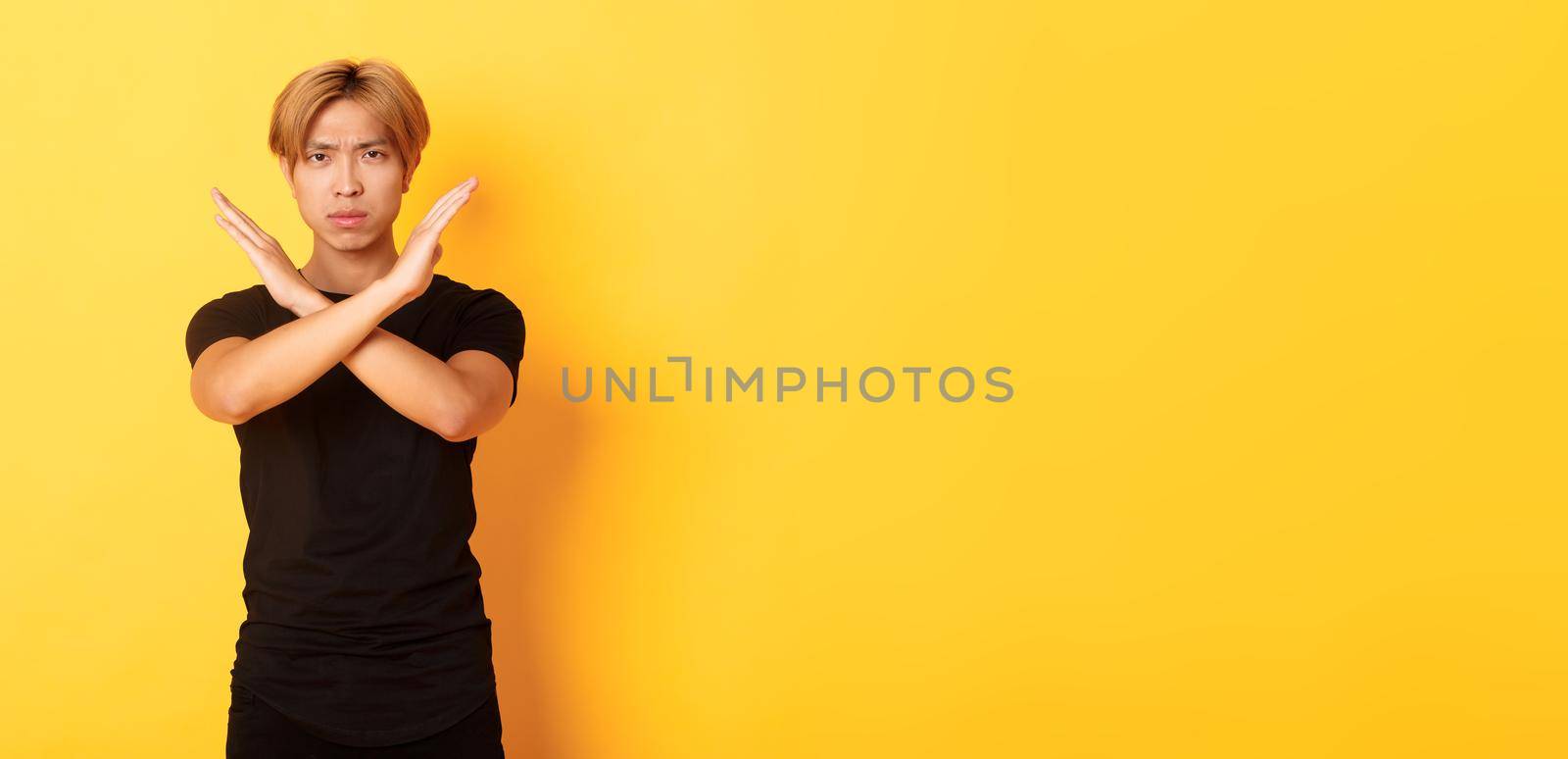 Portrait of disappointed frowning asian man, making cross gesture to stop something bad, standing yellow background by Benzoix