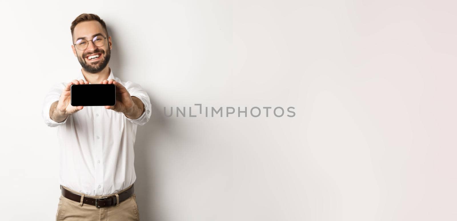 Happy business man showing mobile screen, holding phone horizontally, standing satisfied against white background.