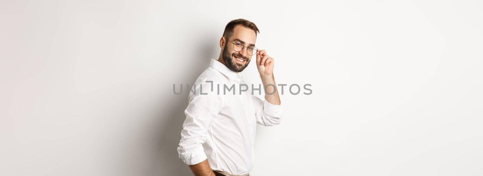 Handsome businessman turn at camera and looking confident, smiling cheeky, standing over white background.
