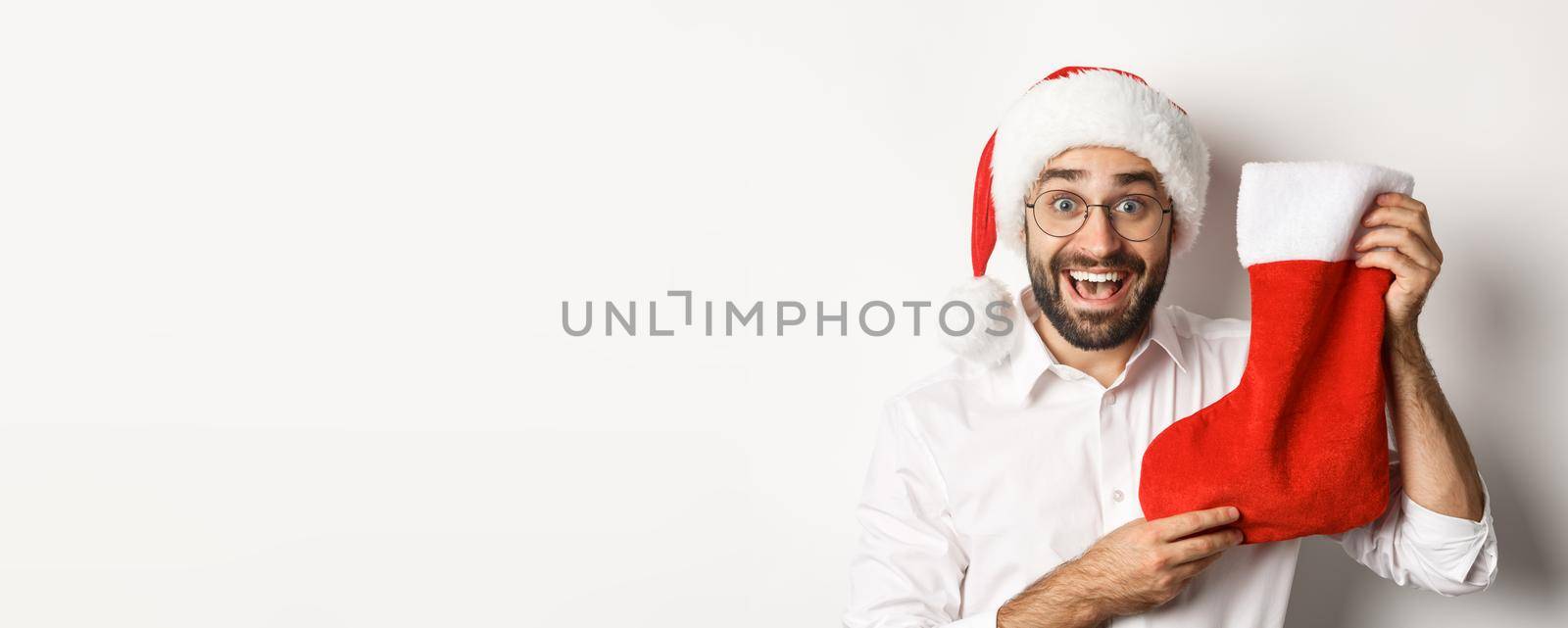 Close-up of happy man celebrating christmas, receive gifts in xmas sock and looking excited, wearing santa hat and glasses, white background.