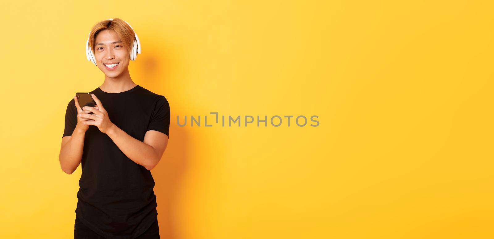 Good-looking smiling, pleased asian guy in wireless headphones, holding smartphone and looking satisfied camera, listening music or podcast.