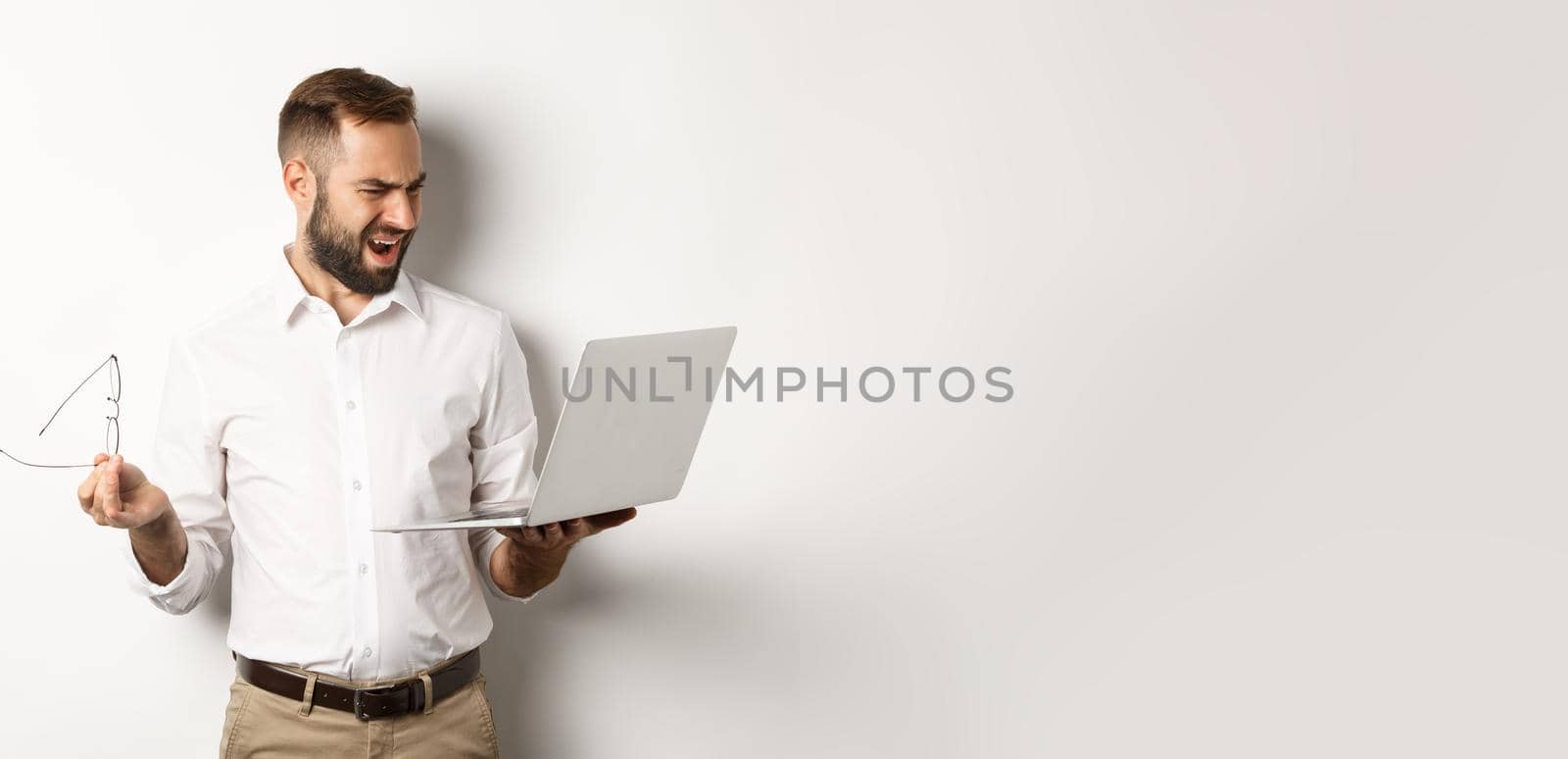 Confused manager looking at laptop, standing disappointed against white background. Copy space