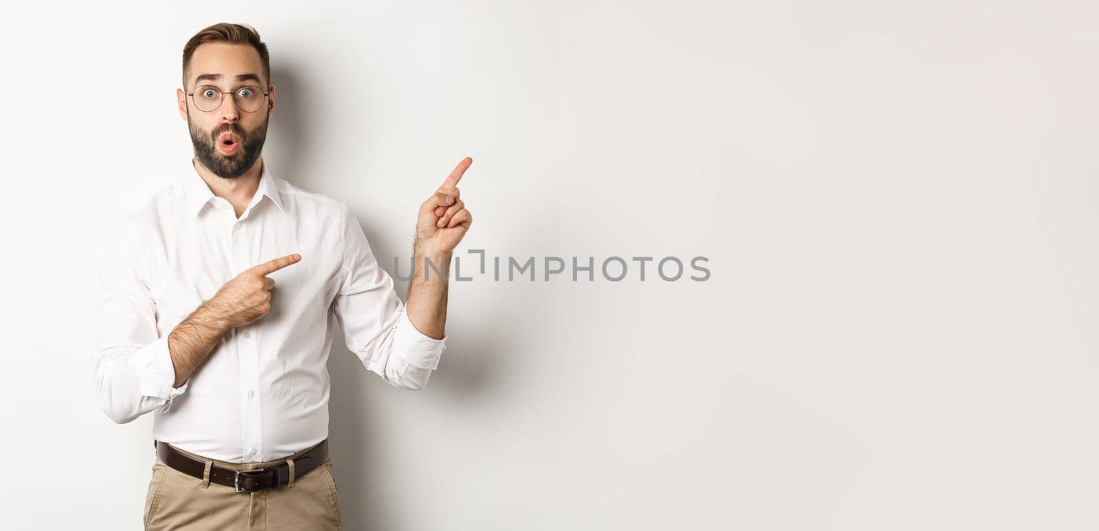 Impressed businessman showing interesting promo offer, pointing fingers right, standing amazed against white background.
