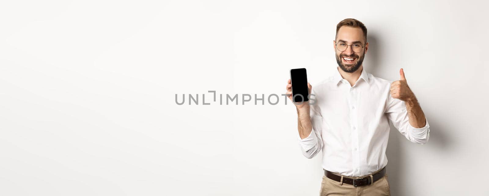 Satisfied business man in glasses showing thumbs up and demonstrating mobile phone screen, recommending app, standing over white background.