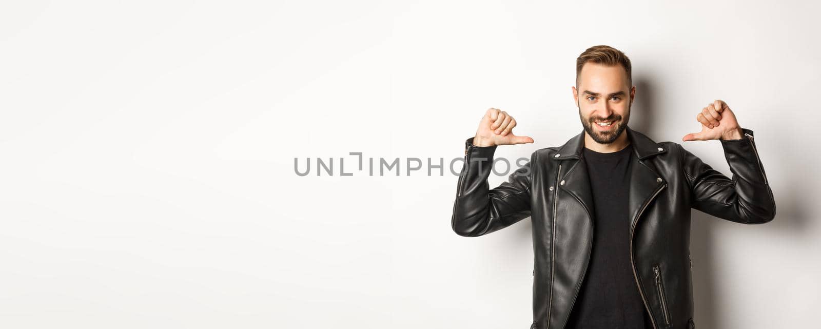 Confident handsome man wearing black leather jacket, pointing at himself and smiling self-assured, standing against white background.