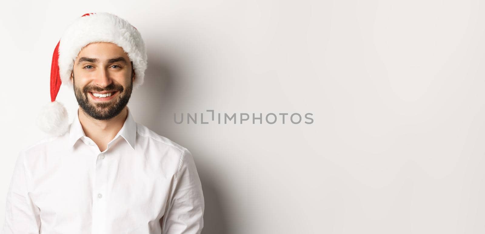 Close-up of happy bearded man celebrating christmas, wearing santa party hat and smiling, standing over white background.