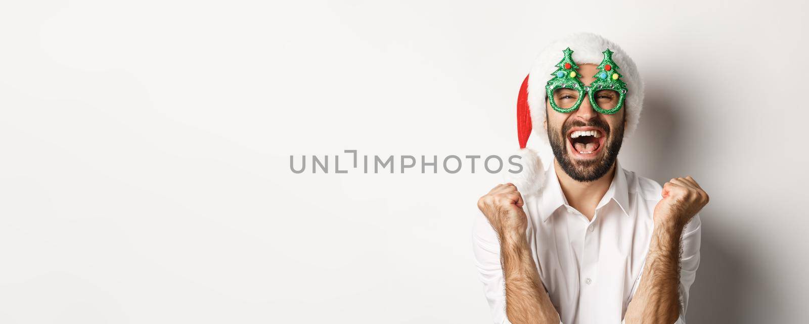 Close-up of man celebrating christmas or new year, wearing xmas party glasses and santa hat, rejoicing and shouting of joy, standing over white background.