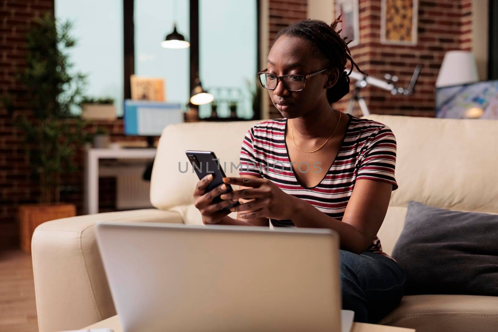 Young african american woman typing message on smartphone, browsing social media, chatting. Freelancer having break, relaxed student in glasses surfing internet on mobile phone