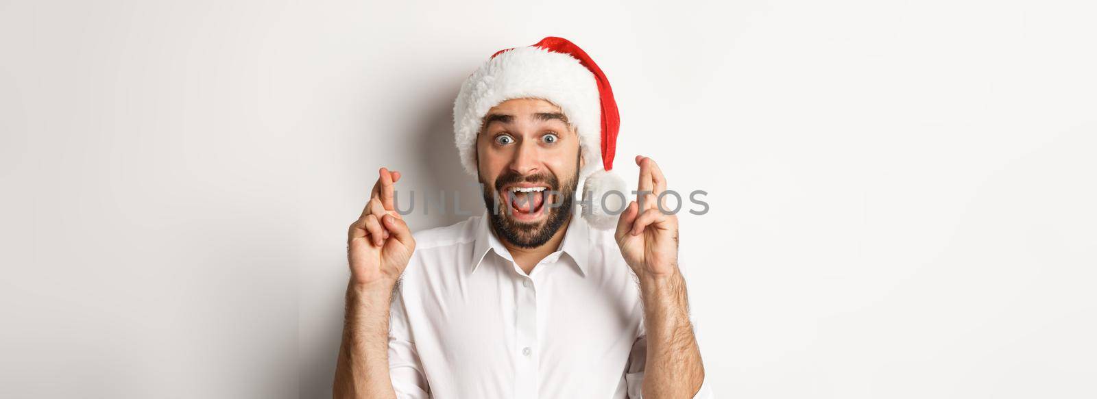 Party, winter holidays and celebration concept. Happy man in santa hat making christmas wish, cross fingers for good luck and looking excited, white background.
