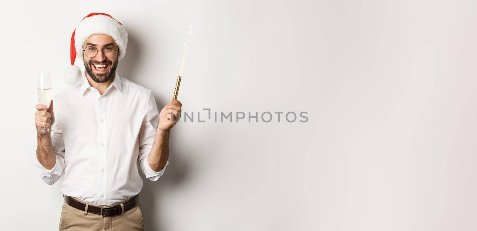 Winter holidays and celebration. Handsome bearded man having New Year party, holding firework sparkle and champagne, wearing santa hat, white background.