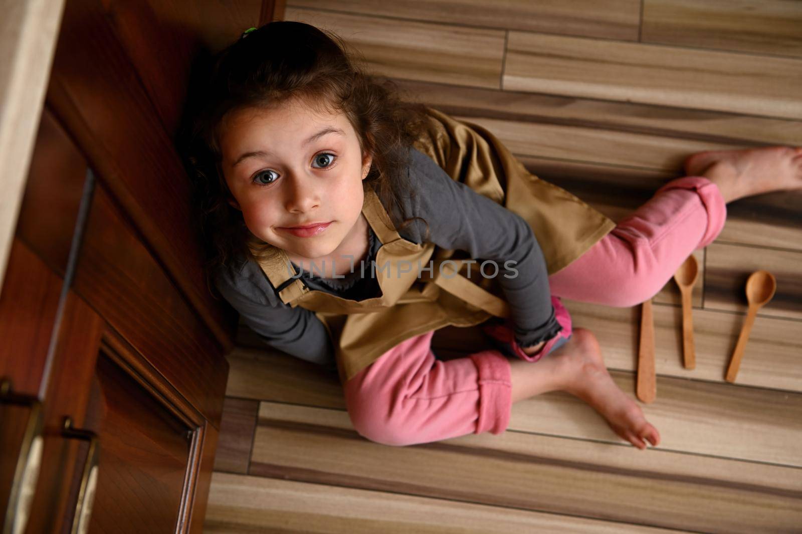 Top view pretty little girl, cute daughter in pink pants, gray long sleeve, beige chef apron and kitchen mittens, sitting barefoot on floor with scattered wooden spoons, looking dreamily up at camera