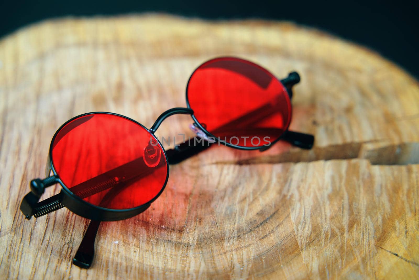 Round sunglasses with red lens on a wooden stand. Fashionable unisex party accessory. Hippie or boho style.