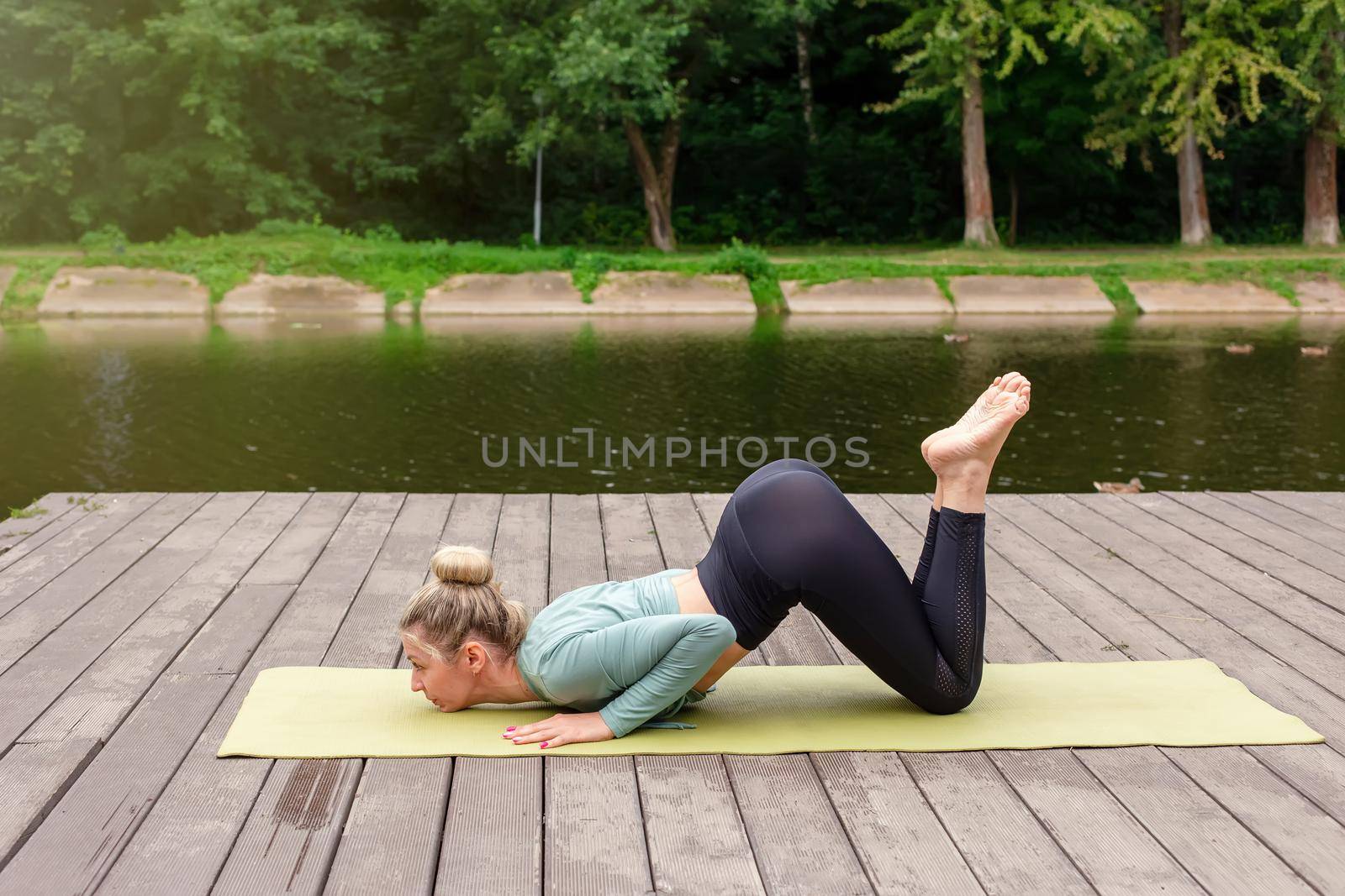 A slender woman in a green top and leggings, in summer, does yoga, stand on a wooden platform by a pond in a park. Copy space.