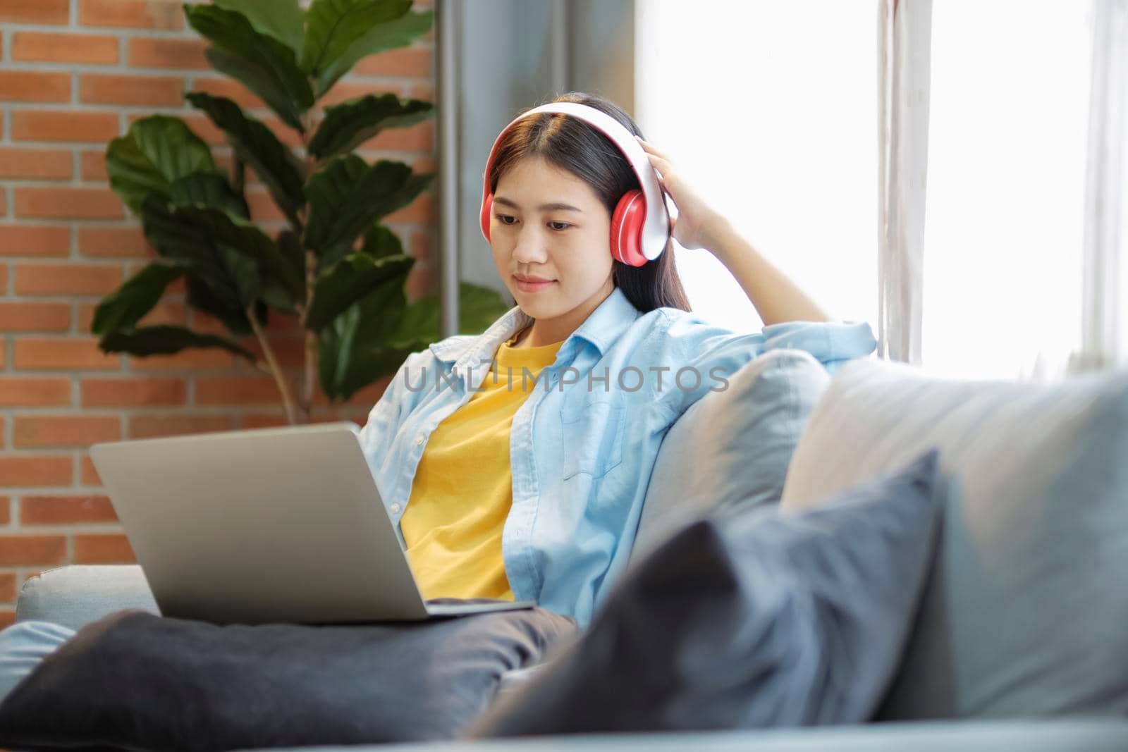 Young asian woman studying, online learing, and listening to music with headset while sitting on couch at home. Listening to podcast, audio book, smiling, and, watching videos using laptop.