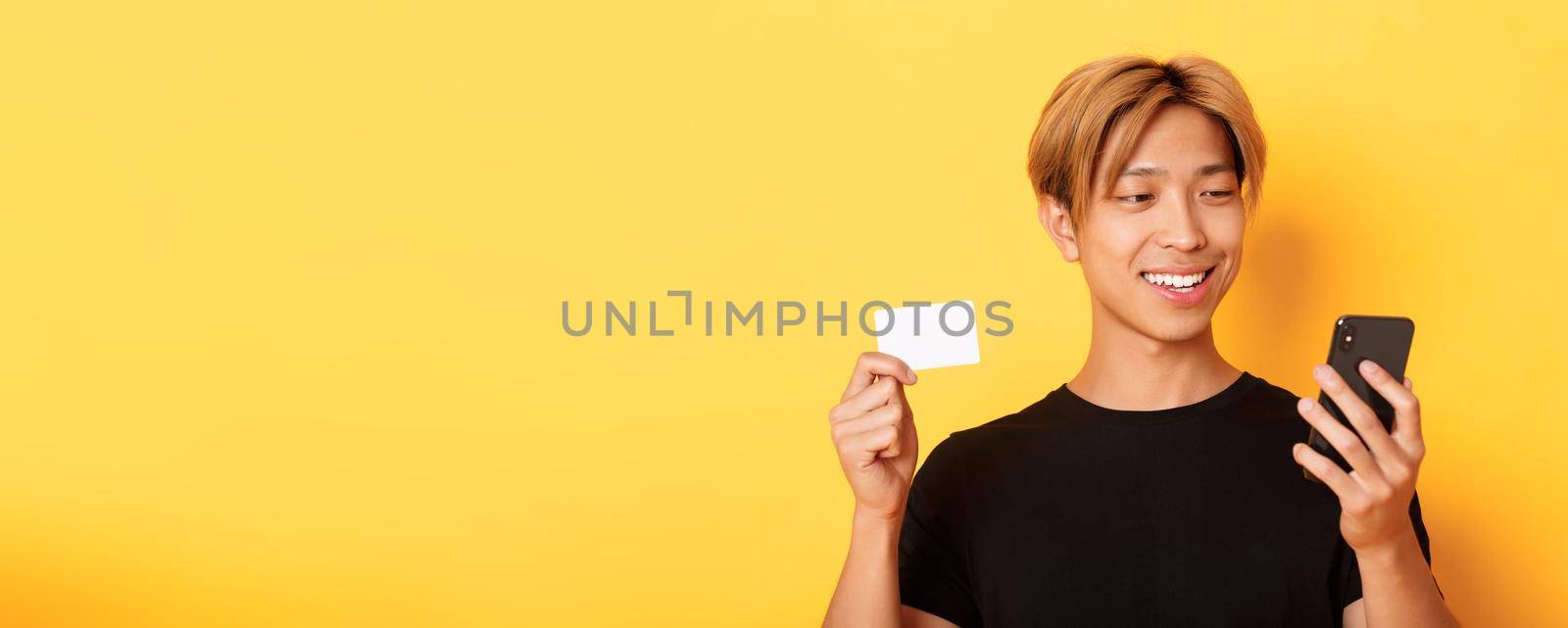 Close-up of handsome stylish asian guy shopping online, looking at mobile phone and smiling, showing credit card, standing over yellow background by Benzoix