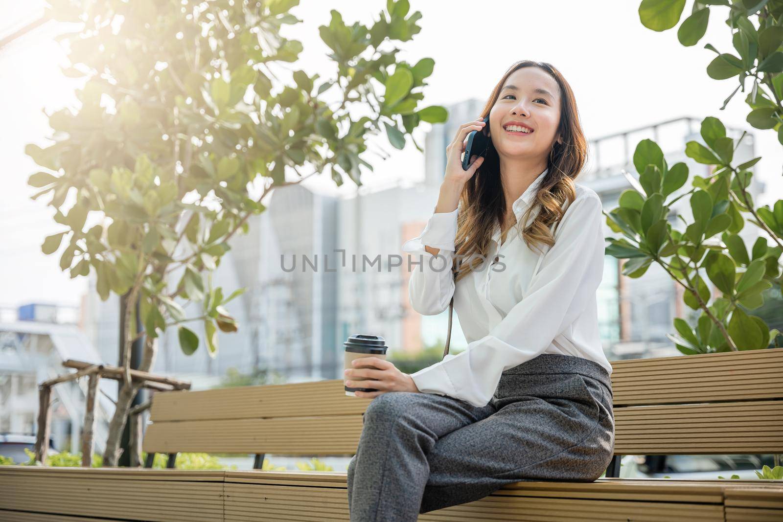 Happy smiling businesswoman sitting talk on mobile phone outside street city near office, Asian business young woman using mobile smartphone talking on cellphone outdoor corporate building exterior