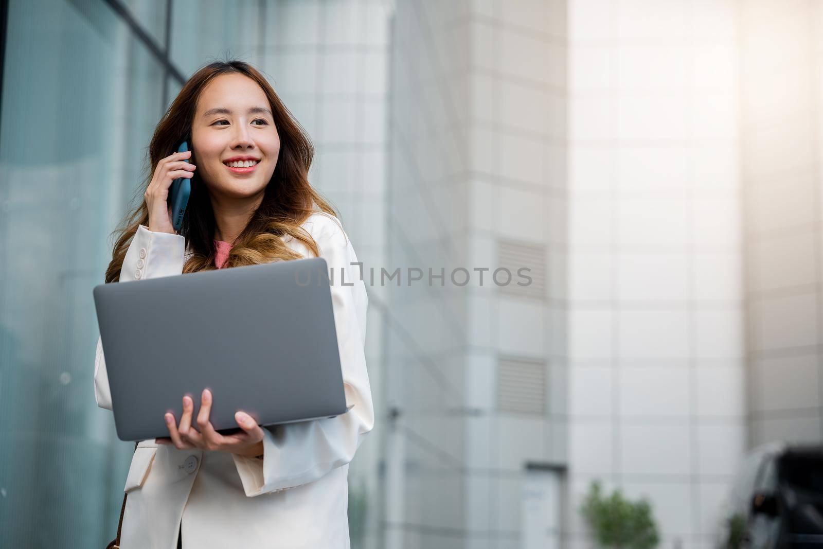 Asian businesswoman working on laptop and talking on cell phone at front building near office by Sorapop