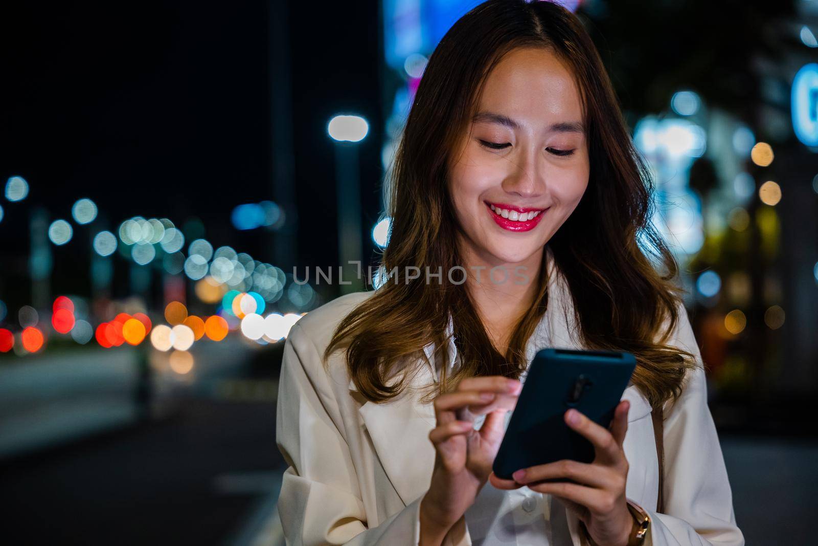 Beautiful young smiling female texting work message on smartphone outside office, Business woman using mobile phone walking through night city street while waiting car to pick up home, social media