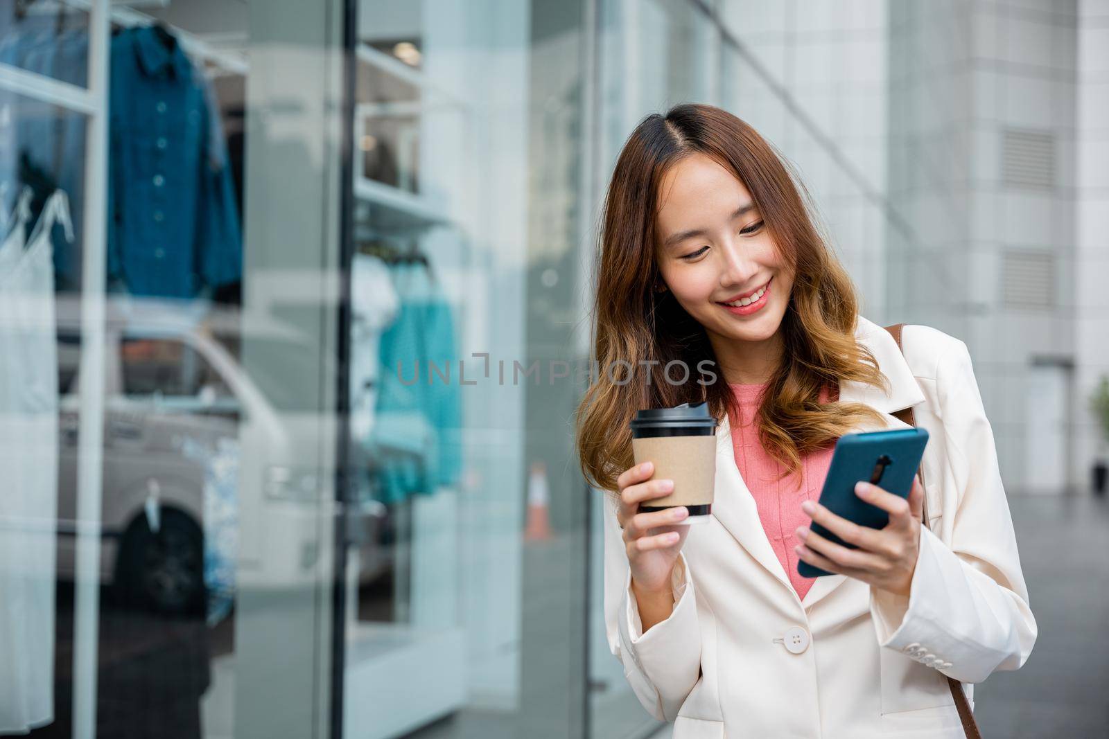 Happy business woman hold mobile phone and paper cup of hot drink outdoor walking on city street, Asian businesswoman holding coffee cup takeaway and smartphone go to work she walking near her office