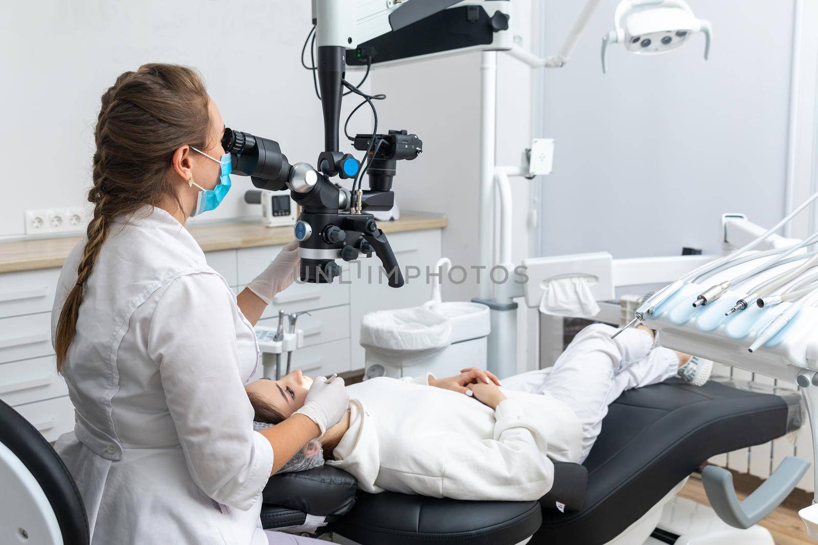 Female dentist using dental microscope treating patient teeth at dental clinic office by Mariakray