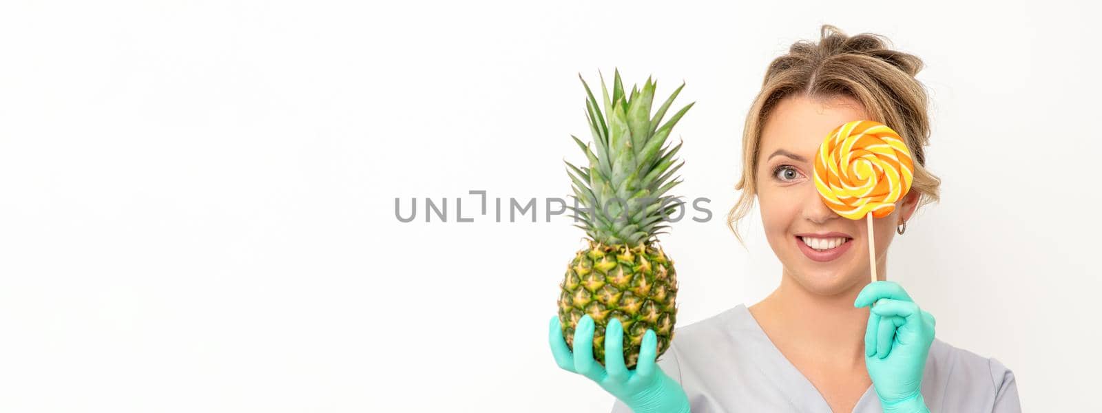 Beautiful smiling beautician holding fresh pineapple and cover her eye with lollipop over white background. Skincare cleansing eco organic. by okskukuruza