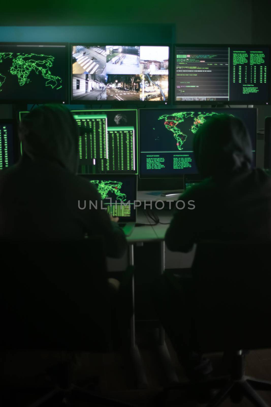 Two hooded hackers hacking security firewall late at night in basement hideout. Vertical image. by Hoverstock