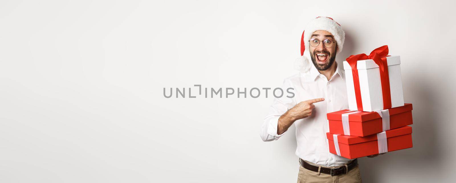 Merry christmas, holidays concept. Surprised man receive xmas gifts, pointing at presents and smiling happy, wearing santa hat, white background.