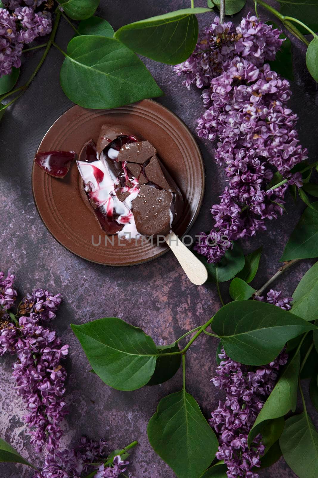 melted raspberry ice cream in chocolate on a wooden stick on a dark burgundy background with lilac flowers, a sweet dessert on a hot spring day, flat lay food. High quality photo