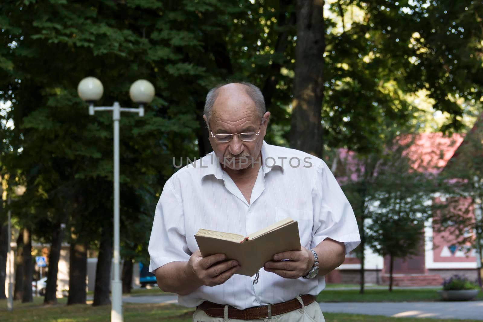 An elderly retired man walks alone in the park. An old, balding man with glasses and a watch on his right hand stands against the background of a red building and reads a book.