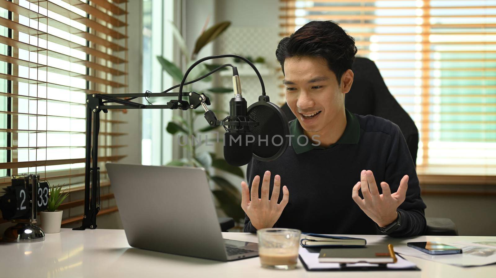 Young podcaster using a microphone and laptop computer recording audio for channel in home studio.