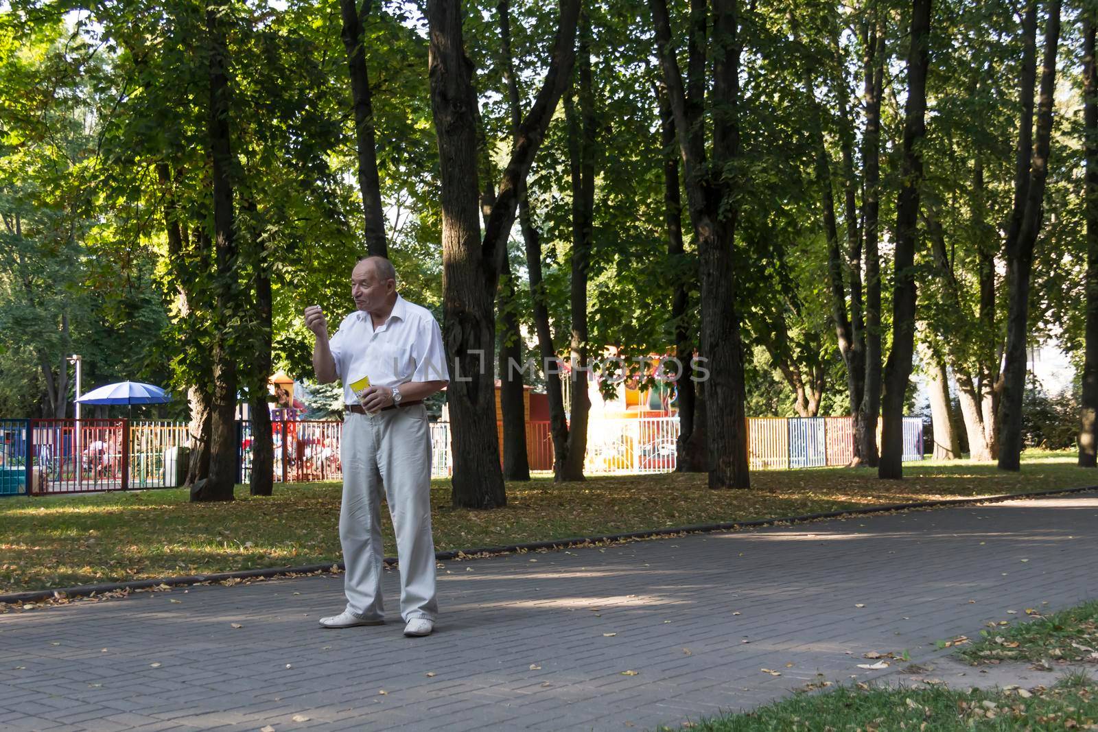 An elderly man walks alone in the park in the summer. A pensioner in a white shirt stands alone in the park with a glass of popcorn.