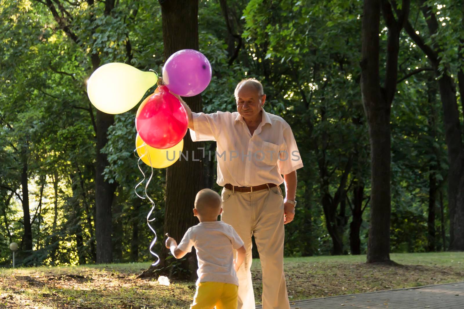 A little boy in yellow shorts and an elderly old man are playing with bright balloons. The family is having fun in the park in the fresh air and celebrating the holiday..