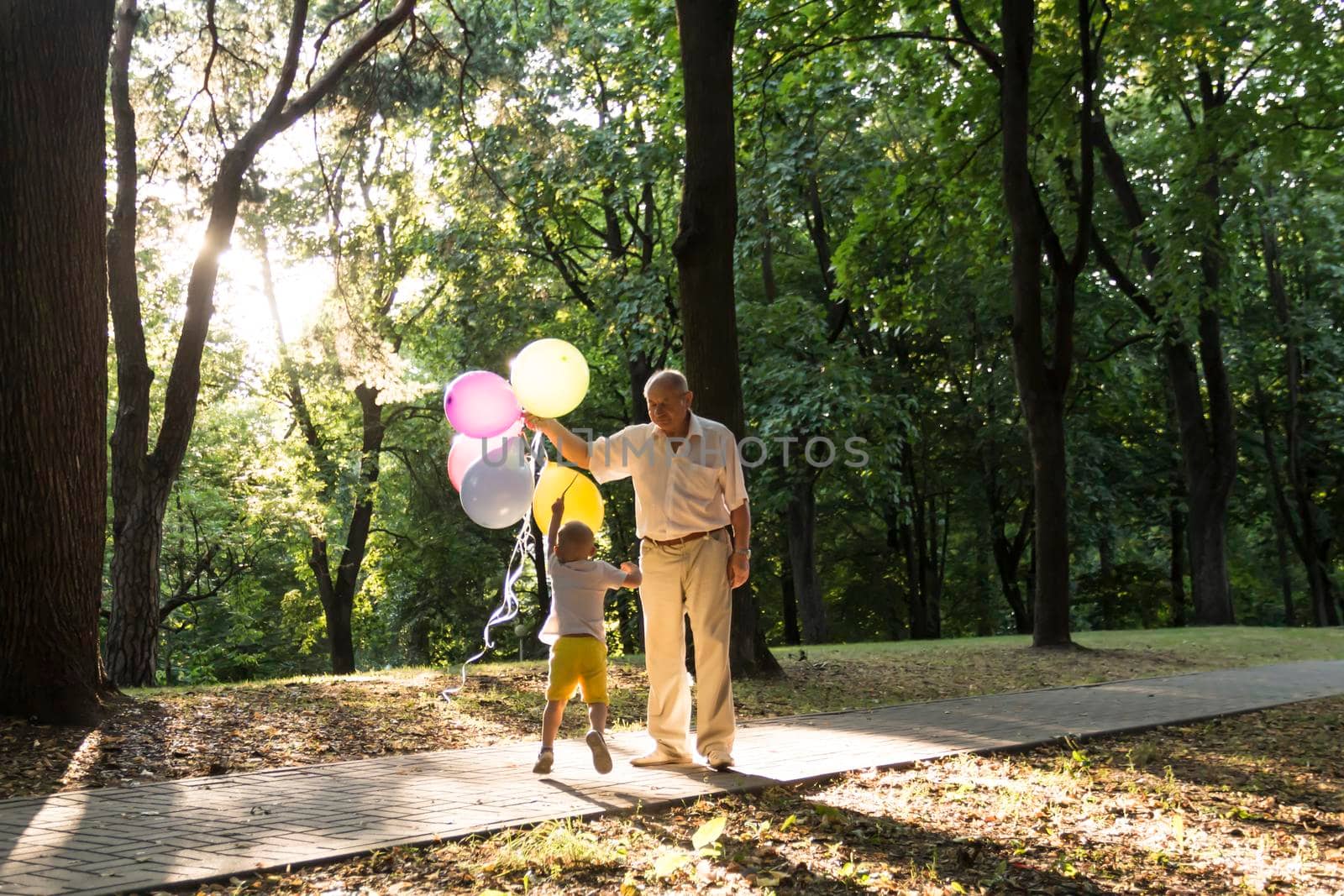 A little boy in yellow shorts and an elderly old man are playing with bright balloons. The family is having fun in the park in the fresh air and celebrating the holiday..